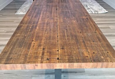 A long wooden dining table with a rustic finish in a modern living room. The room features wood flooring, two large speakers, a TV on the wall, and a leather sofa set on a textured rug.