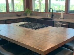 A spacious kitchen with a large wooden island featuring a built-in stovetop. There are cushioned bar stools around the island. Large windows provide a view of trees and greenery outside, creating a bright and inviting space.