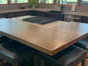 A spacious kitchen with a large wooden island featuring a built-in stovetop. There are cushioned bar stools around the island. Large windows provide a view of trees and greenery outside, creating a bright and inviting space.