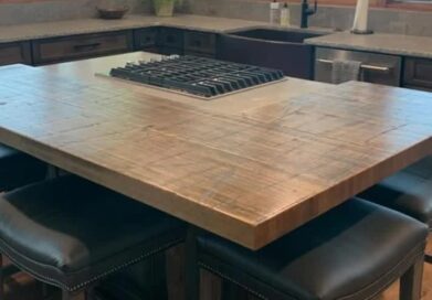 A spacious kitchen with a large wooden island featuring a built-in stovetop. There are cushioned bar stools around the island. Large windows provide a view of trees and greenery outside, creating a bright and inviting space.