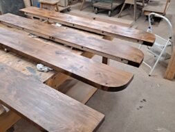 Wooden planks are laid out on a workbench in a workshop, ready for assembly or construction. The smooth, polished surfaces reveal a rich brown color, enhanced by the overhead lighting. Various tools and equipment are visible in the background.