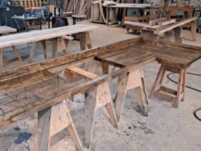 A woodworking shop with several wooden tables supported by saw horses. Wood shavings are scattered on the floor. Various tools and wooden planks are visible in the background.