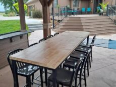 A patio with a long wooden dining table surrounded by black chairs. Steps lead up to another outdoor area with turquoise chairs. The setting is under an awning, with grass and a house visible in the background.