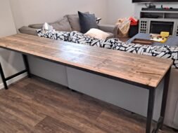 Wooden table with black metal legs positioned behind a gray sectional sofa in a living room. The sofa has patterned black and white cushions, and the room has wooden floors, a wall-mounted TV, and a small cabinet.