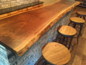 A Live Edge Tops wooden bar counter with a natural finish is set against a brick wall. Four round wooden stools with metal frames are neatly aligned along the bar, which sits atop a floor made of wood planks.