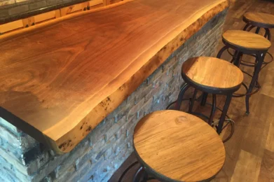 A Live Edge Tops wooden bar counter with a natural finish is set against a brick wall. Four round wooden stools with metal frames are neatly aligned along the bar, which sits atop a floor made of wood planks.