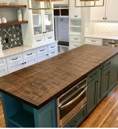 A modern kitchen with a long wooden island countertop. The island has teal cabinets and stainless steel appliances. Surrounding white cabinetry features open shelves, a black and white decorative backsplash, and a tile backsplash near the sink.