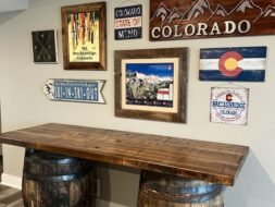 A rustic wooden table made from barrels sits beneath a wall decorated with various Colorado-themed signs and artwork, including mountains, a state flag, and ski-themed pieces. The decor is set against a light-colored wall.