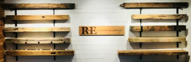 A display of rustic wooden shelves and mantels crafted from reclaimed wood, mounted on a white wall, features a central sign reading RE Reclaimed Enterprises. The Reclaimed Wood Mantels add a rugged, natural aesthetic to the setting.