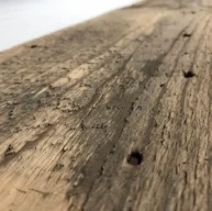 Close-up of a piece from the Reclaimed Wood Mantels collection, showcasing its weathered wooden surface with visible grain, small holes, and a rough texture that evokes the charm of antique mantels. The reclaimed wood has a rustic, aged appearance and stretches diagonally across the image against a blurred backdrop.