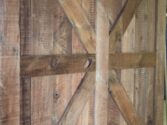 A rustic wooden barn door with a natural finish, featuring a prominent X-shaped pattern made from wide planks. The door showcases a variety of wood textures and knots, highlighting its handcrafted appearance.