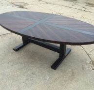 A large, oval wooden table with a dark finish and a cross pattern on the tabletop, situated on a rough, cracked concrete surface. The table has a sturdy black pedestal base with two wide legs.