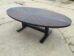 A large, oval wooden table with a dark finish and a cross pattern on the tabletop, situated on a rough, cracked concrete surface. The table has a sturdy black pedestal base with two wide legs.