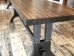 A long, rectangular wooden table with sturdy metal legs stands on a rustic hardwood floor. The tables smooth surface showcases the wood grain, contrasting with the industrial-style legs underneath. Another table is visible in the background.