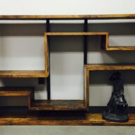 A wooden, geometrically designed bookcase with staggered shelves stands against a white wall. A small black statue of a cowboy is placed on the bottom right shelf, adding a decorative touch to the minimalist furniture piece.
