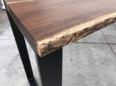 Close-up of a wooden table corner made from dark-stained wood with a natural live edge design, showcasing textured wood grain and unique markings. The table stands on black metal legs, set against a concrete floor background.