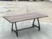 A wooden table with a smooth, rectangular top and natural edges stands on a metal frame with four wheels, placed on a concrete surface. The background is an open outdoor area.