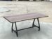 A wooden table with a smooth, rectangular top and natural edges stands on a metal frame with four wheels, placed on a concrete surface. The background is an open outdoor area.