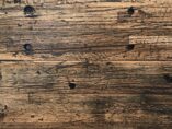 A close-up of a textured wooden surface with visible knots and grain patterns, displaying shades of brown and dark streaks, suggesting an aged or reclaimed wood look.