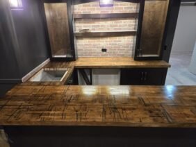 A home bar with a rustic wooden countertop and brick backsplash. Cabinets with glass doors sit above the counter. The area is dimly lit by hanging lights, creating a cozy atmosphere.