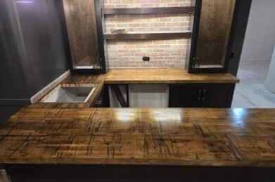 A home bar with a rustic wooden countertop and brick backsplash. Cabinets with glass doors sit above the counter. The area is dimly lit by hanging lights, creating a cozy atmosphere.