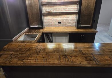 A home bar with a rustic wooden countertop and brick backsplash. Cabinets with glass doors sit above the counter. The area is dimly lit by hanging lights, creating a cozy atmosphere.