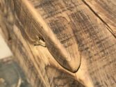 Close-up of a piece of aged, weathered wood with visible grain and a prominent crack. The wood surface shows various textures and patterns, highlighted by natural lighting.