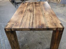 A handcrafted wooden table with a rustic finish stands in a workshop, surrounded by tools, workbenches, and equipment. The table features thick legs and a polished, textured tabletop with visible wood grain and knots.