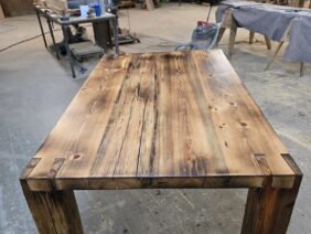 A handcrafted wooden table with a rustic finish stands in a workshop, surrounded by tools, workbenches, and equipment. The table features thick legs and a polished, textured tabletop with visible wood grain and knots.
