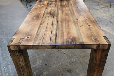 A handcrafted wooden table with a rustic finish stands in a workshop, surrounded by tools, workbenches, and equipment. The table features thick legs and a polished, textured tabletop with visible wood grain and knots.