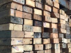 A stack of rough, weathered wooden beams piled high in a warehouse. The timber exhibits natural variations in color and age, with visible knots and grain patterns. The background shows part of a concrete wall.
