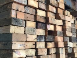 A stack of rough, weathered wooden beams piled high in a warehouse. The timber exhibits natural variations in color and age, with visible knots and grain patterns. The background shows part of a concrete wall.