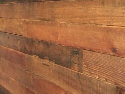 Close-up of a rustic wooden wall with staggered horizontal planks in various shades of brown. The surface shows natural grain and texture, with a small section of a red window frame visible at the top left corner.