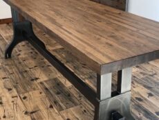 A wooden table with a dark metal frame stands on a rustic wooden floor. The tabletop has a rich wood grain texture, and the metal supports feature a sleek, industrial design.