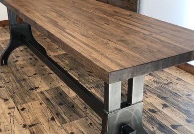 A wooden table with a dark metal frame stands on a rustic wooden floor. The tabletop has a rich wood grain texture, and the metal supports feature a sleek, industrial design.