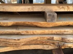 Stacked slabs of raw, unfinished wood with irregular, natural edges are neatly arranged. The wood appears to be in a workshop setting, hinting at carpentry or woodworking projects.