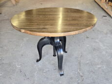 A round wooden table with a dark polished top and black metal legs stands on a concrete floor in a woodworking shop with tools and wooden planks in the background.