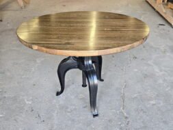 A round wooden table with a dark polished top and black metal legs stands on a concrete floor in a woodworking shop with tools and wooden planks in the background.
