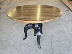 A round wooden table with a dark polished top and black metal legs stands on a concrete floor in a woodworking shop with tools and wooden planks in the background.