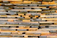 A close-up view of a neatly stacked pile of wooden planks, varying in color from light to dark brown. The planks are arranged horizontally, showing their textured surfaces and edges.