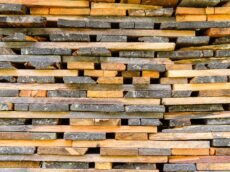 A close-up view of a neatly stacked pile of wooden planks, varying in color from light to dark brown. The planks are arranged horizontally, showing their textured surfaces and edges.