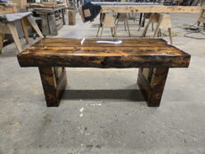 A handmade wooden bench with a thick, glossy, polished top and sturdy legs sits in a workshop surrounded by tools, wood pieces, and workbenches.