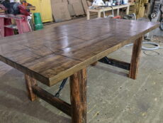 A large, rustic wooden table with thick legs and a patterned top sits in a workshop surrounded by tools, supplies, and industrial equipment.