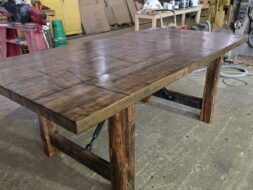 A large, rustic wooden table with thick legs and a patterned top sits in a workshop surrounded by tools, supplies, and industrial equipment.