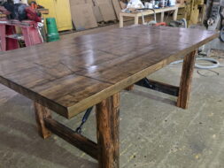 A large, rustic wooden table with thick legs and a patterned top sits in a workshop surrounded by tools, supplies, and industrial equipment.