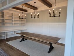 A spacious room featuring a rustic wooden table on a light rug, open white built-in shelves along the wall, two black metal chandeliers, wood ceiling beams, and white paneled walls.