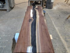 A long wooden table with a glossy finish and a dark, wavy resin river running through its center sits on sawhorses in a workshop filled with tools and wood materials.