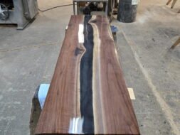 A long wooden table with a glossy finish and a dark, wavy resin river running through its center sits on sawhorses in a workshop filled with tools and wood materials.