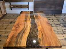 A polished wooden table with a black resin river running through the center stands on a wooden floor, with samples of wood panels displayed on the wall behind it.