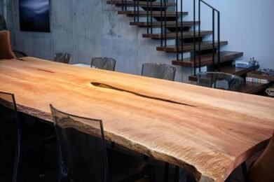 A modern dining area features a large wooden table with a natural edge. Above, an artistic chandelier with numerous hanging lights adds elegance. A staircase with dark railings ascends to a second level, and large windows provide ample natural light.